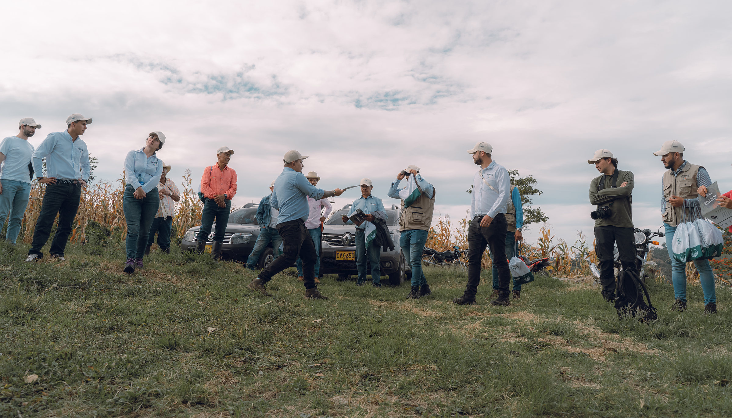 The field team and growers gathered on a hilltop at the edge of a maize plot: one of the team explaining with a cut stem in his hand, others taking notes and a photographer recording the session
