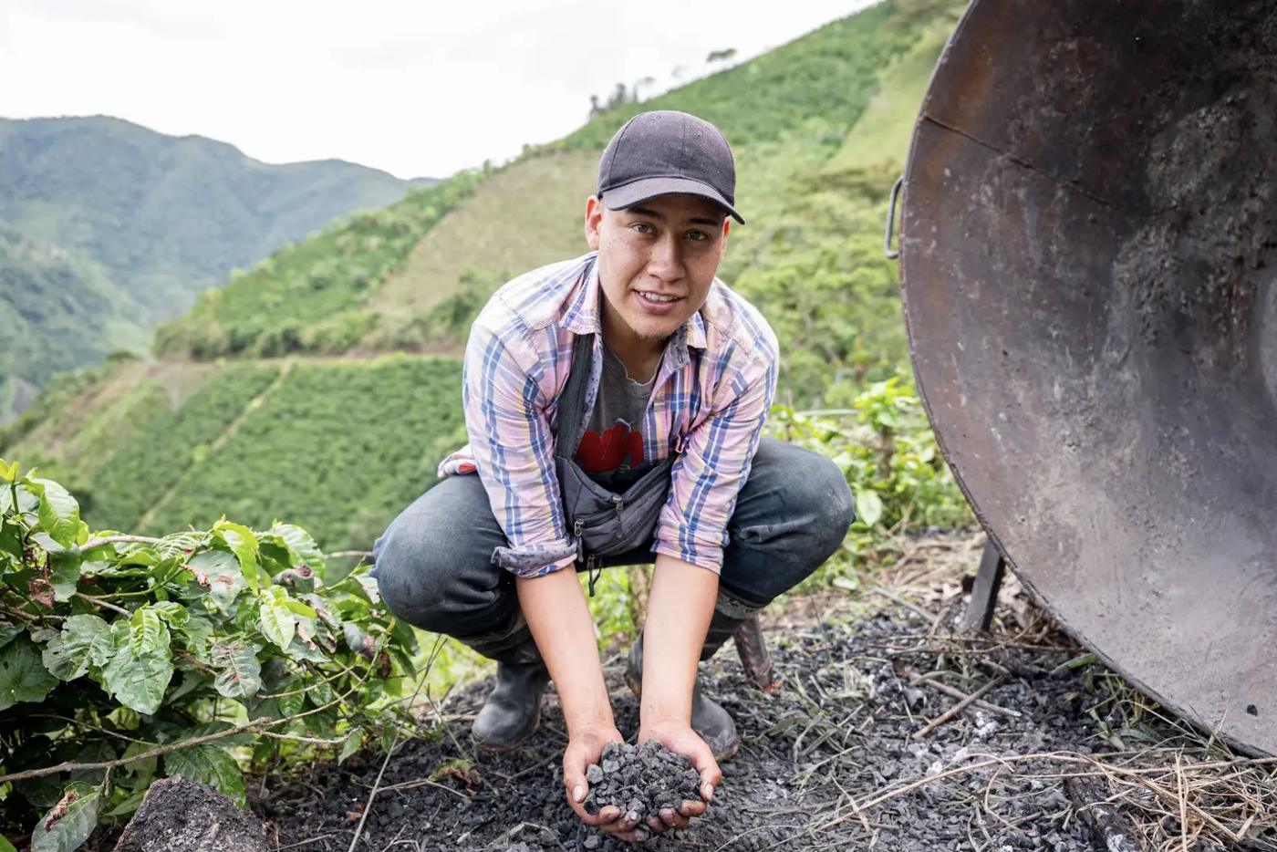 Daniel Alexander Perdomo crouching on his farm, holding a handful of biochar made from his own coffee prunings, the kiln beside him